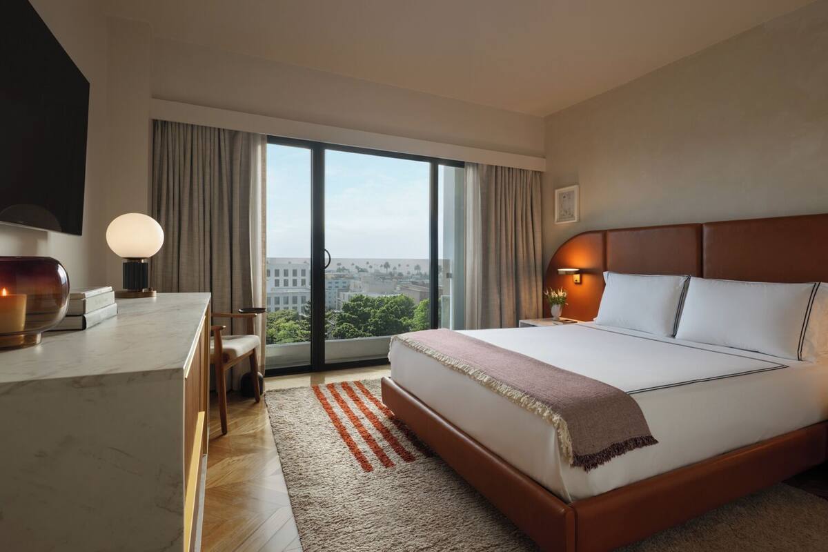 A modern hotel room with a large bed, a desk with a lamp, a television, and a view of the city through large windows.
