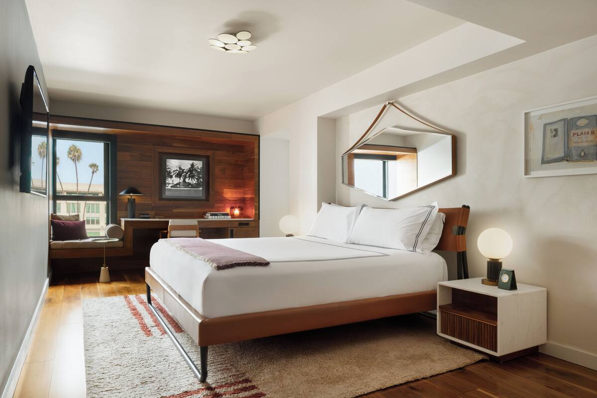 A modern bedroom with a large bed, bedside tables, a mirror, and a view of palm trees through the window.