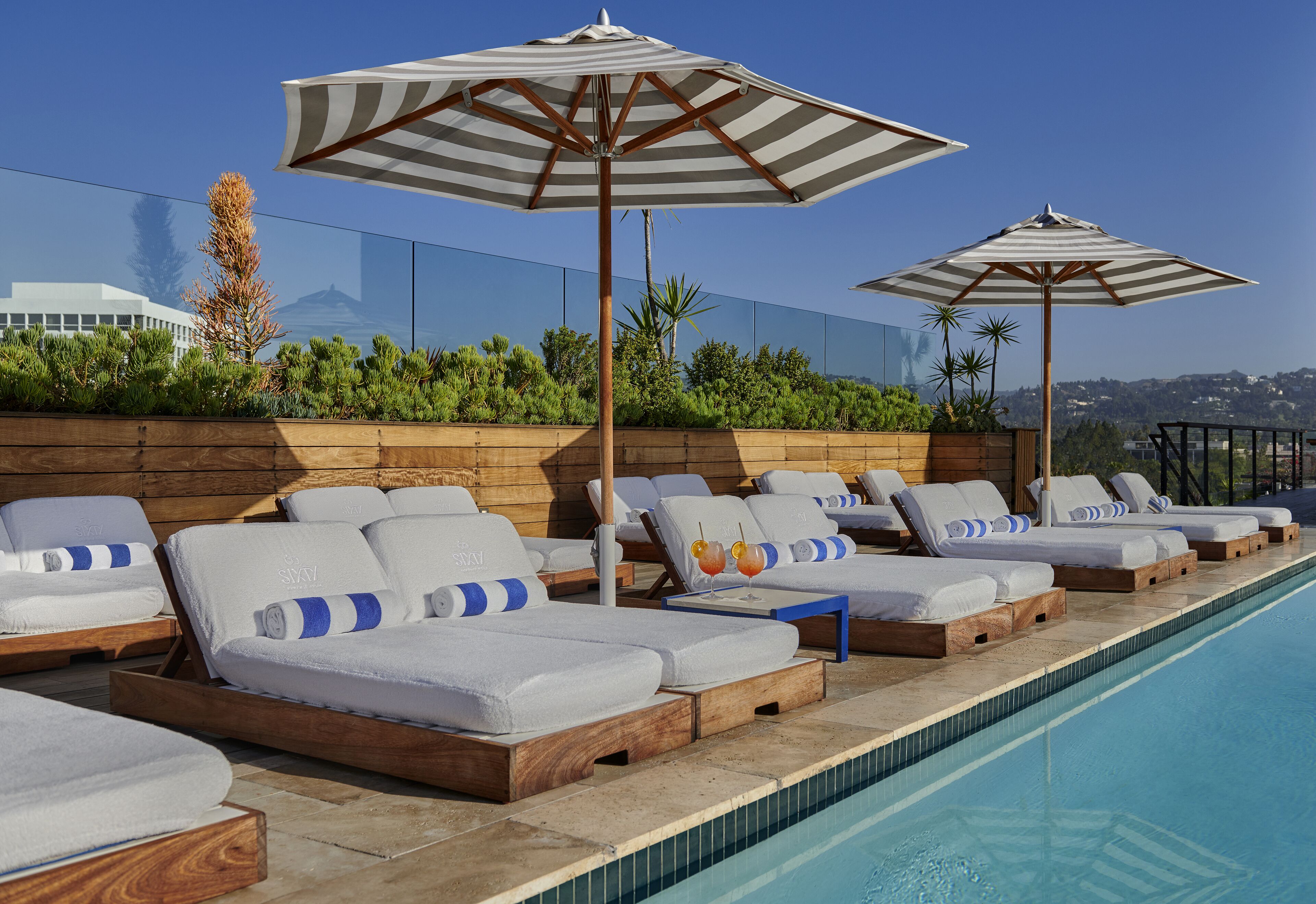 Zona junto a la piscina con tumbonas y sombrillas, una terraza de madera y una vista despejada del horizonte de la ciudad.