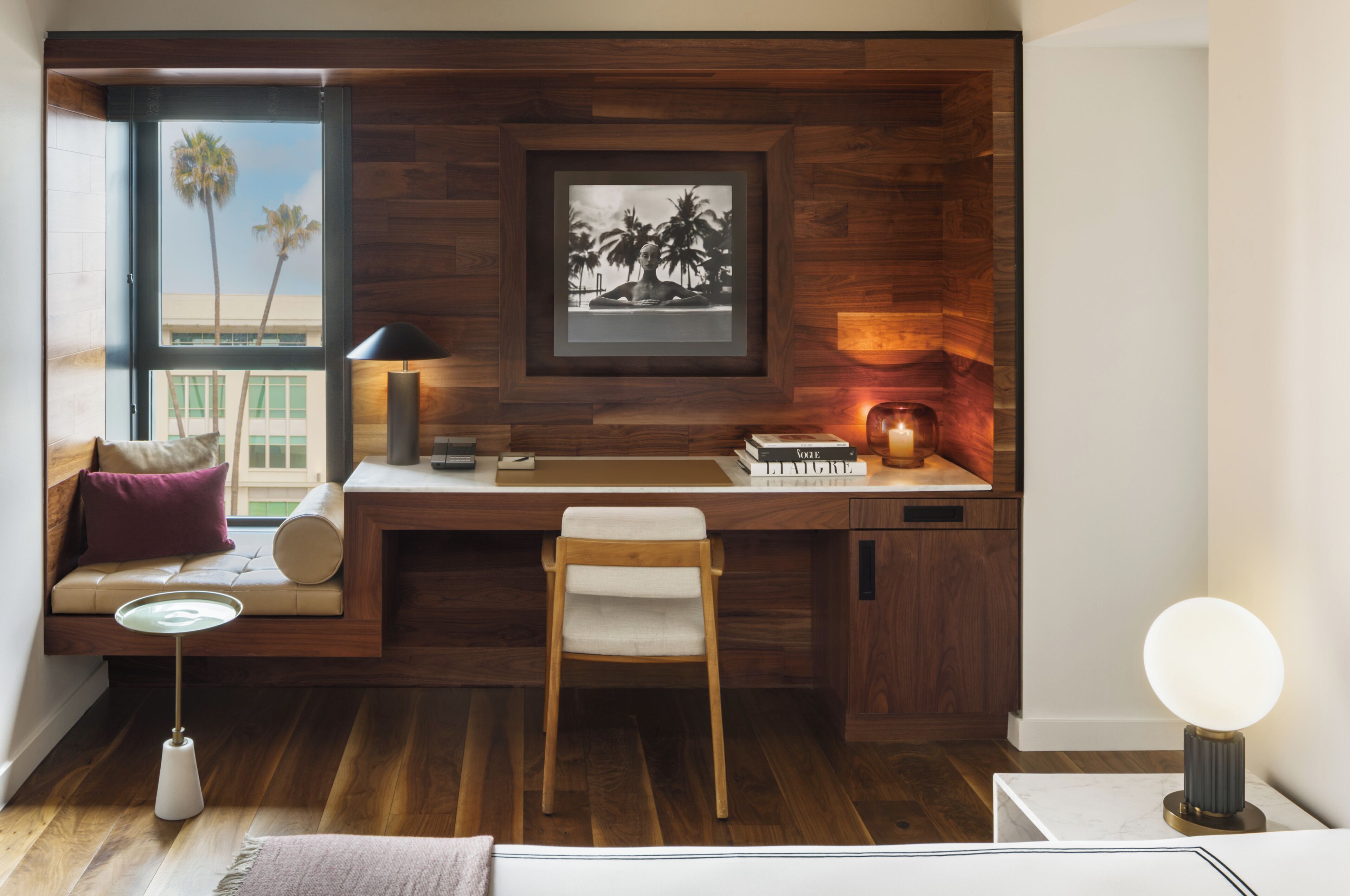 A modern workspace with a wooden desk, a chair, a window with a view of palm trees, and a framed picture on the wall.