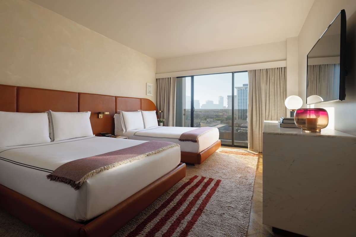 A hotel room with two beds, a large window with a city view, a flat-screen TV, and a desk with a lamp.