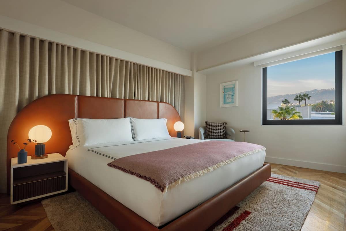A modern hotel room with a large bed, two bedside tables with lamps, a view of palm trees, and a cityscape through the window.