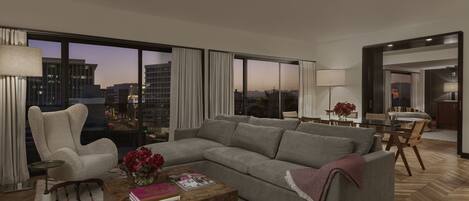 A modern living room with a large grey sectional sofa, a wooden coffee table, and a city view through floor-to-ceiling windows.