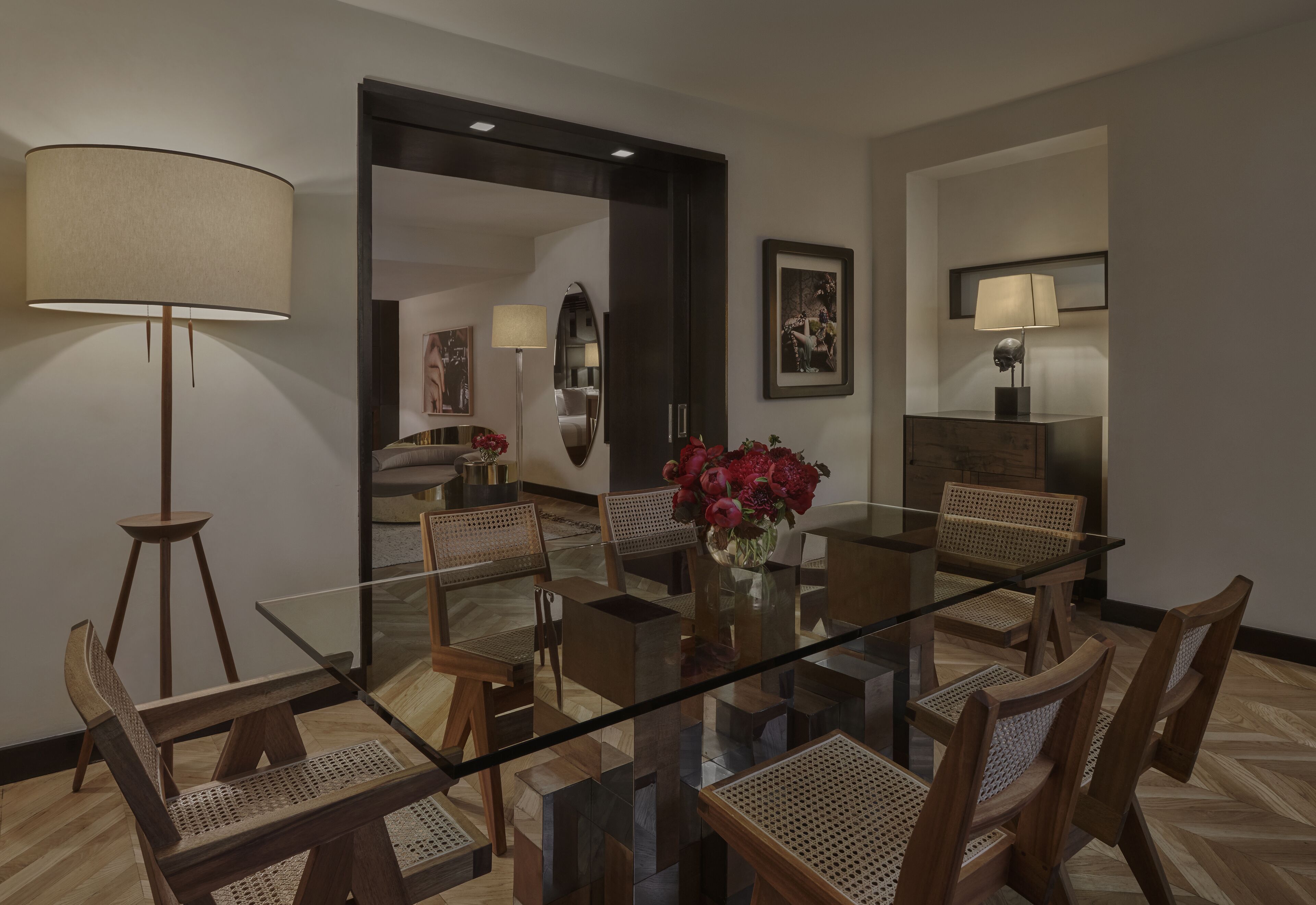 Un comedor con una mesa de vidrio rodeada de sillas de madera, un florero con flores y dos lámparas de pie.