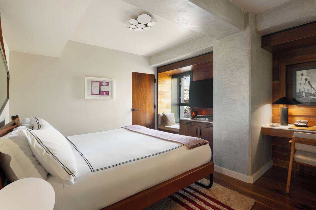 A bedroom with a bed, a desk, and a chair. There is a window with a view of the outside.