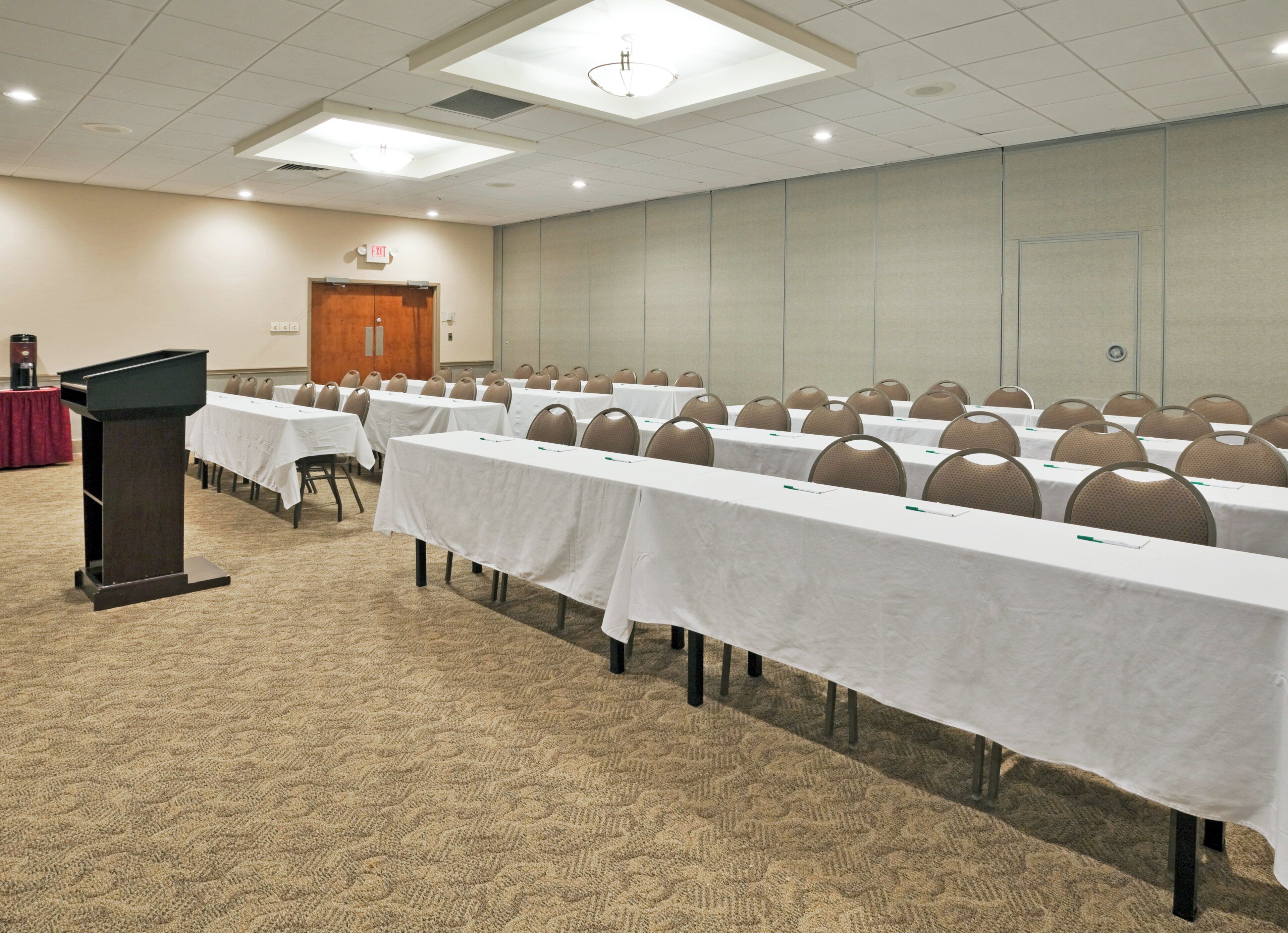 A conference room with rows of tables and chairs, a podium, and an exit door.