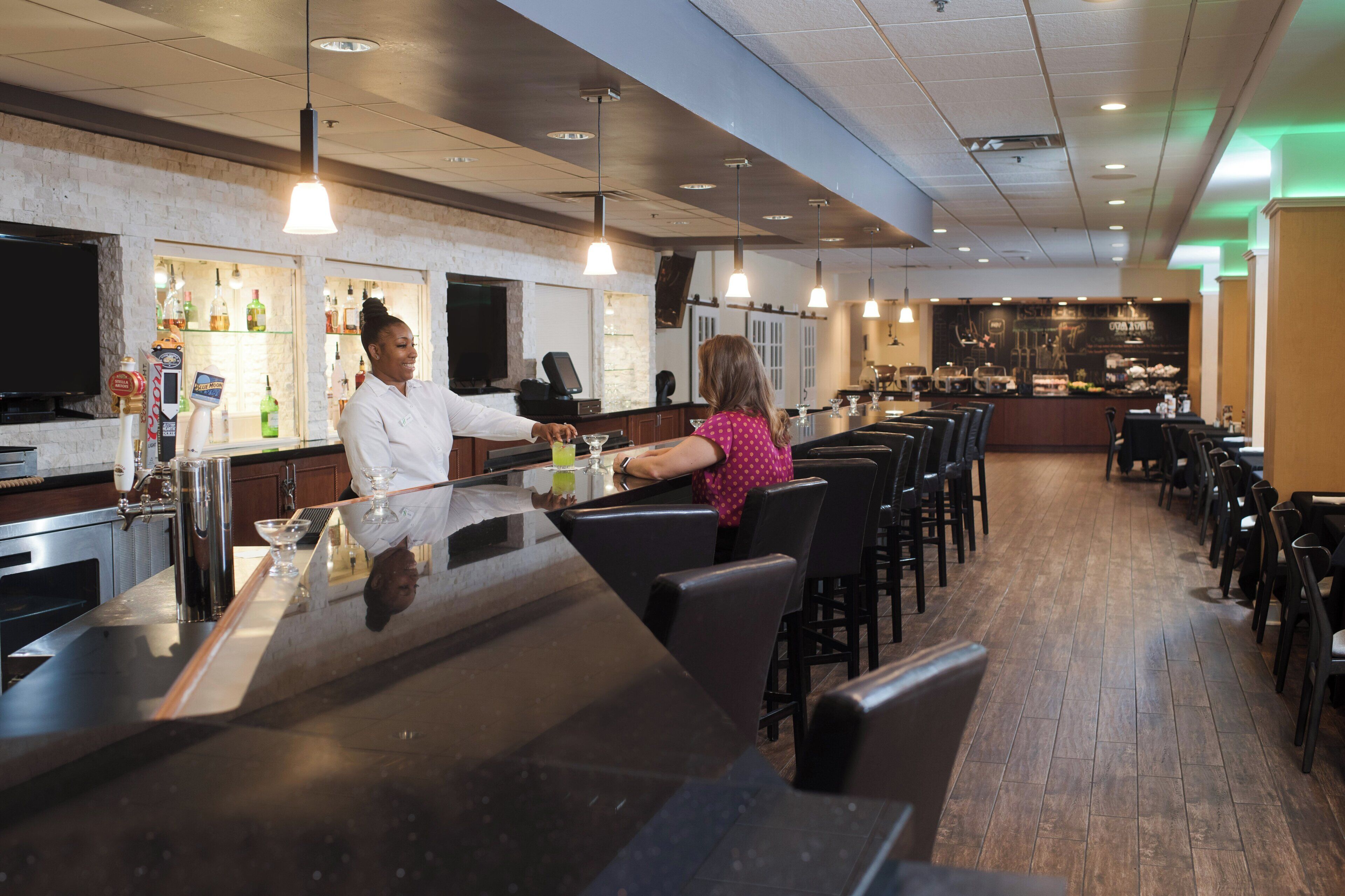 A bar area with a bartender serving a customer, a television mounted on the wall, and a variety of bottles on shelves.