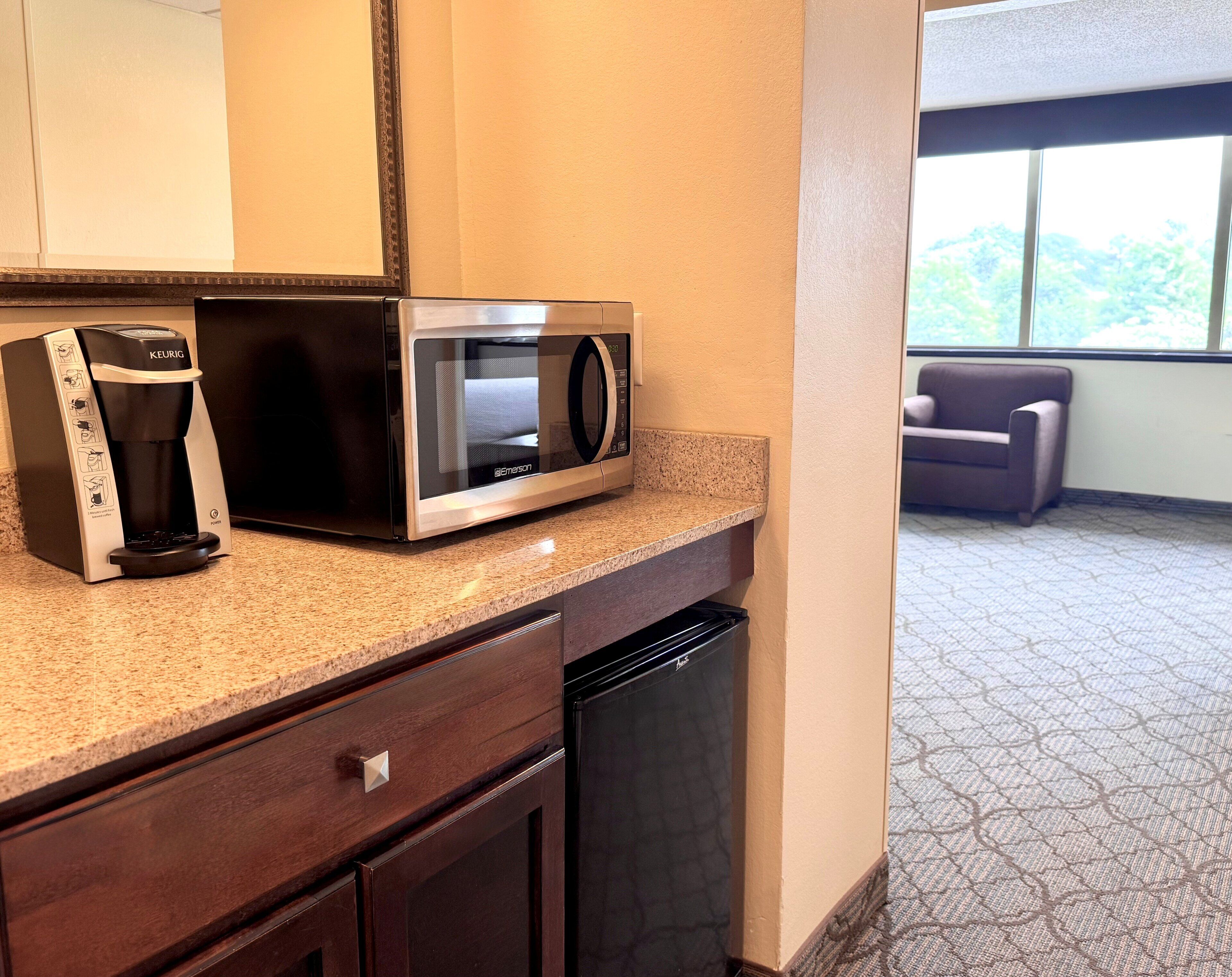 A kitchenette with a Keurig coffee maker, a microwave, a dishwasher, and a window with a view of trees.