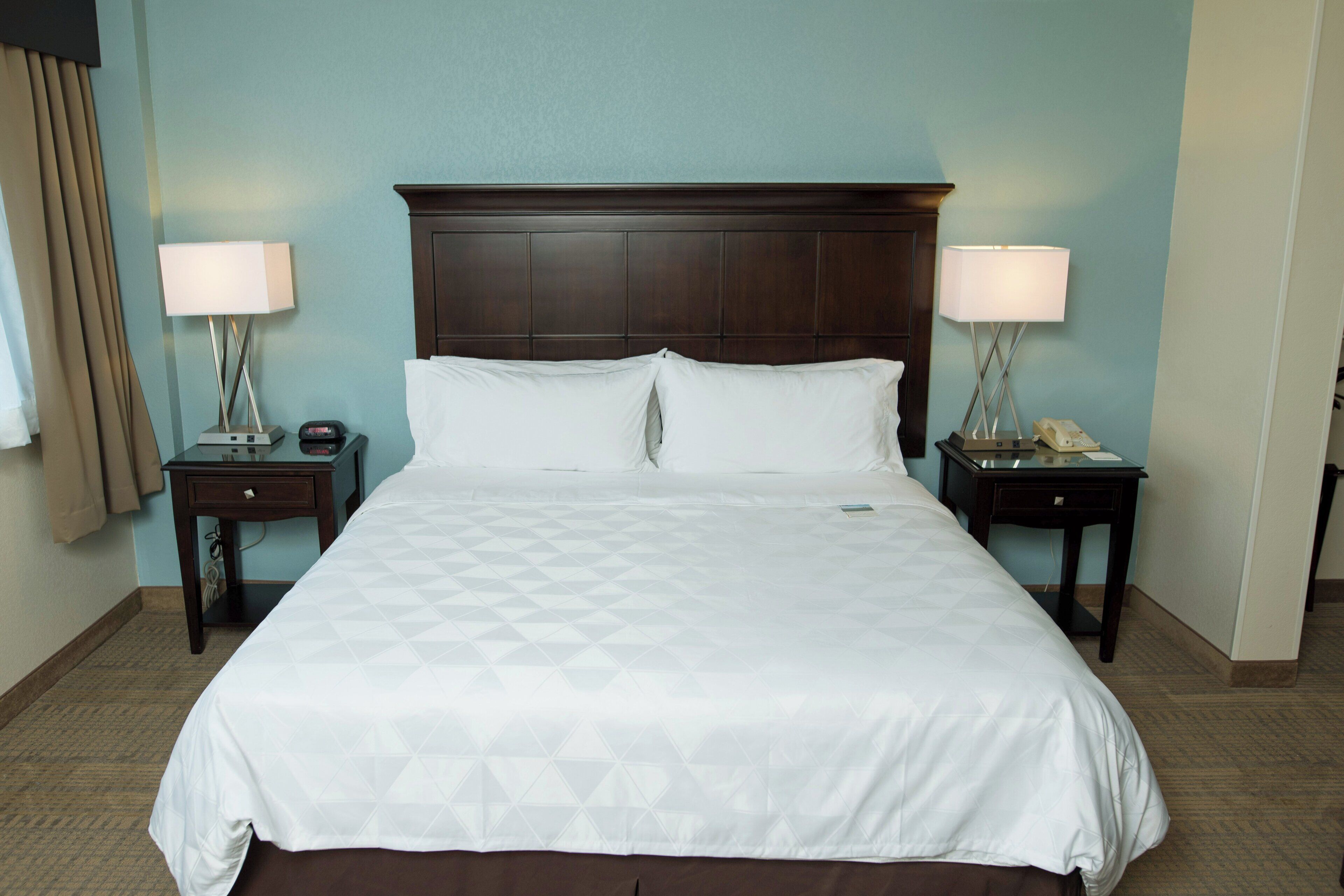 A hotel room with a large bed, two bedside tables, a nightstand, a wooden headboard, and a wall-mounted lamp.