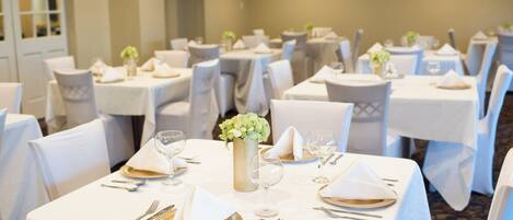 Un salón de banquetes con mesas redondas dispuestas para comer, cada una con mantel blanco, cubiertos y cristalería.
