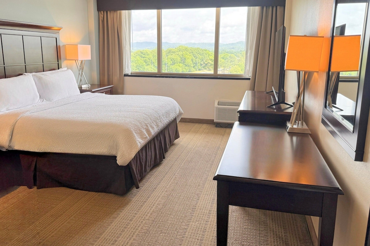 A hotel room with a bed, a desk, a chair, a lamp, a window with a view, and a mirror.