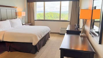 A hotel room with a bed, a desk, a chair, a lamp, a window with a view, and a mirror.