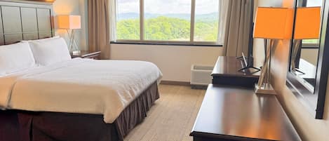 Ett hotellrum med en säng, ett skrivbord, en stol, en lampa, ett fönster med utsikt och en spegel.