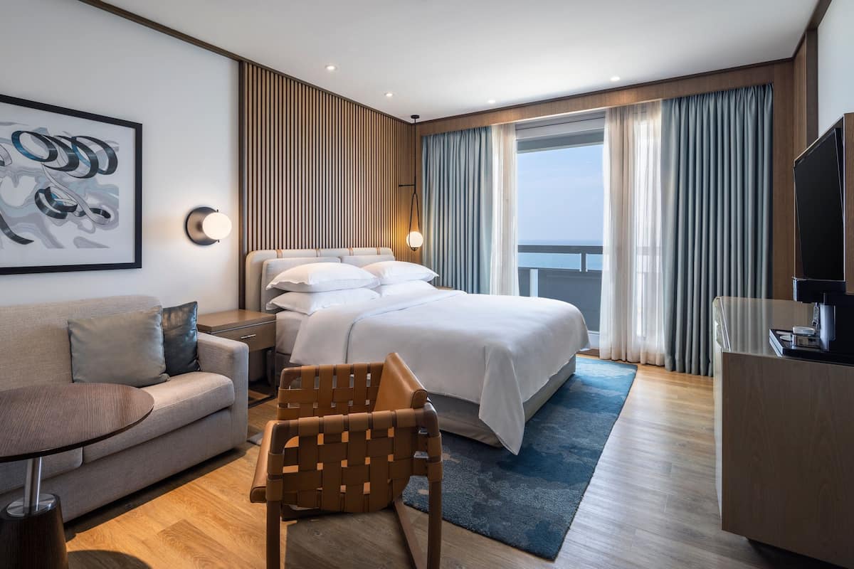 Una habitación de hotel moderna con una cama, un sofá, una silla, un escritorio, un televisor y un amplio ventanal con cortinas.