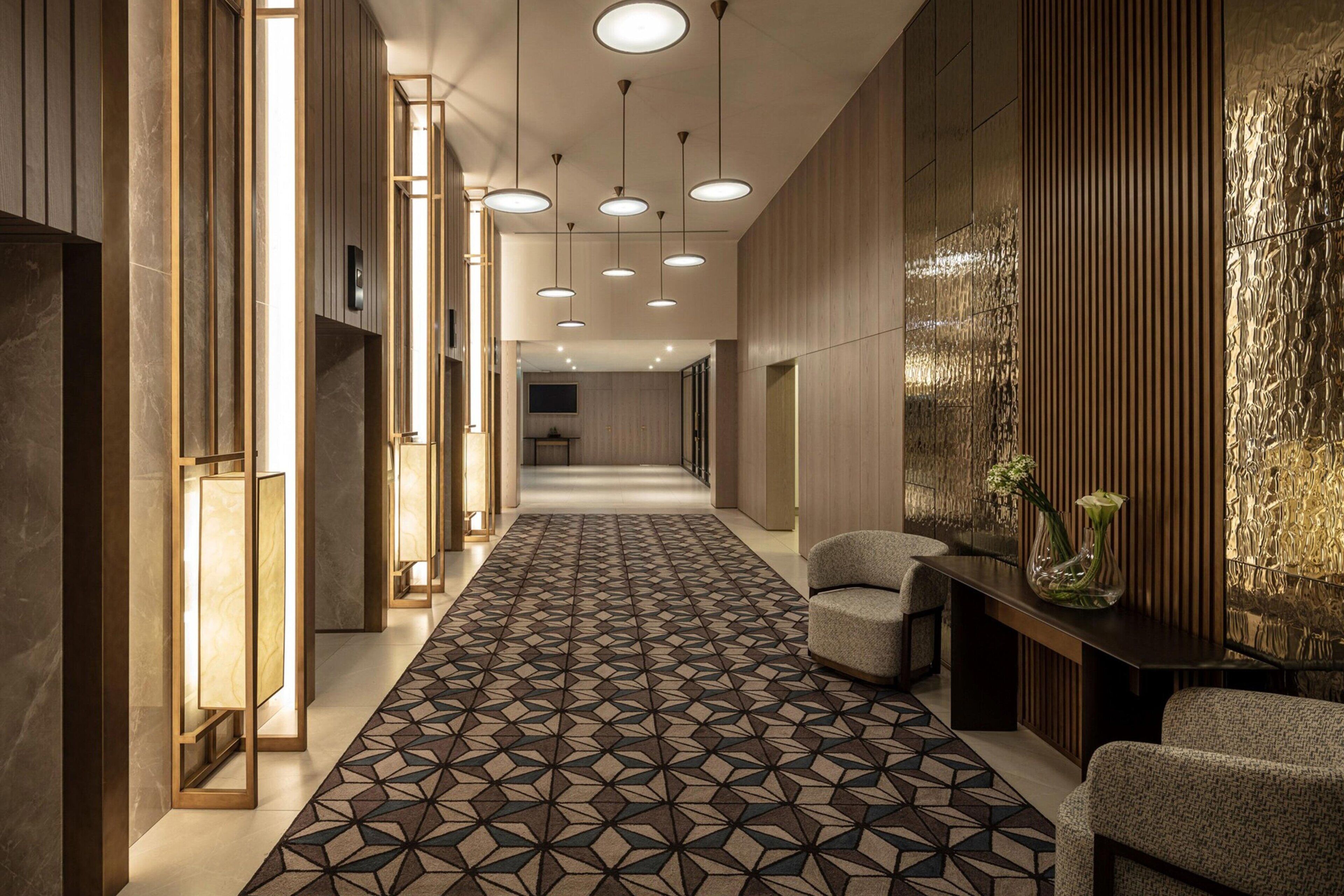 Un hall d’hôtel moderne avec un tapis à motifs géométriques, des luminaires suspendus et un comptoir d’accueil doté de chaises.