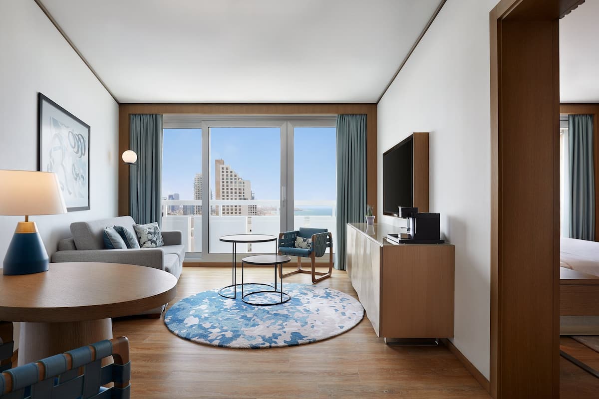 Una habitación de hotel moderna con sofá, mesa de comedor y sillas, una pequeña cocina, televisión de pantalla plana y un amplio ventanal con vistas a la ciudad.