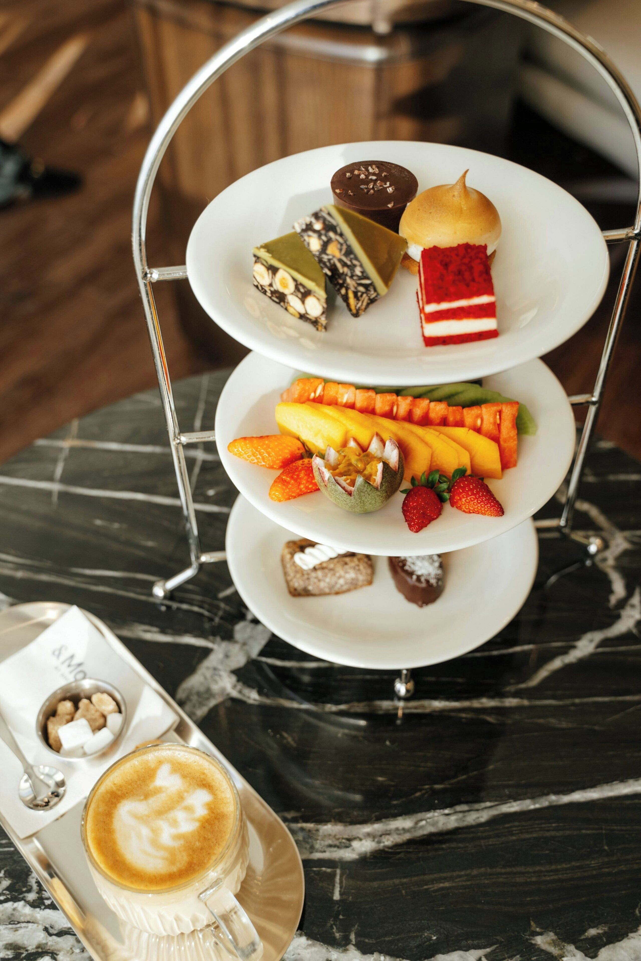 Un soporte de tres niveles con diversos dulces y frutas, una taza de café y un cuenco pequeño de nueces sobre una mesa de mármol.