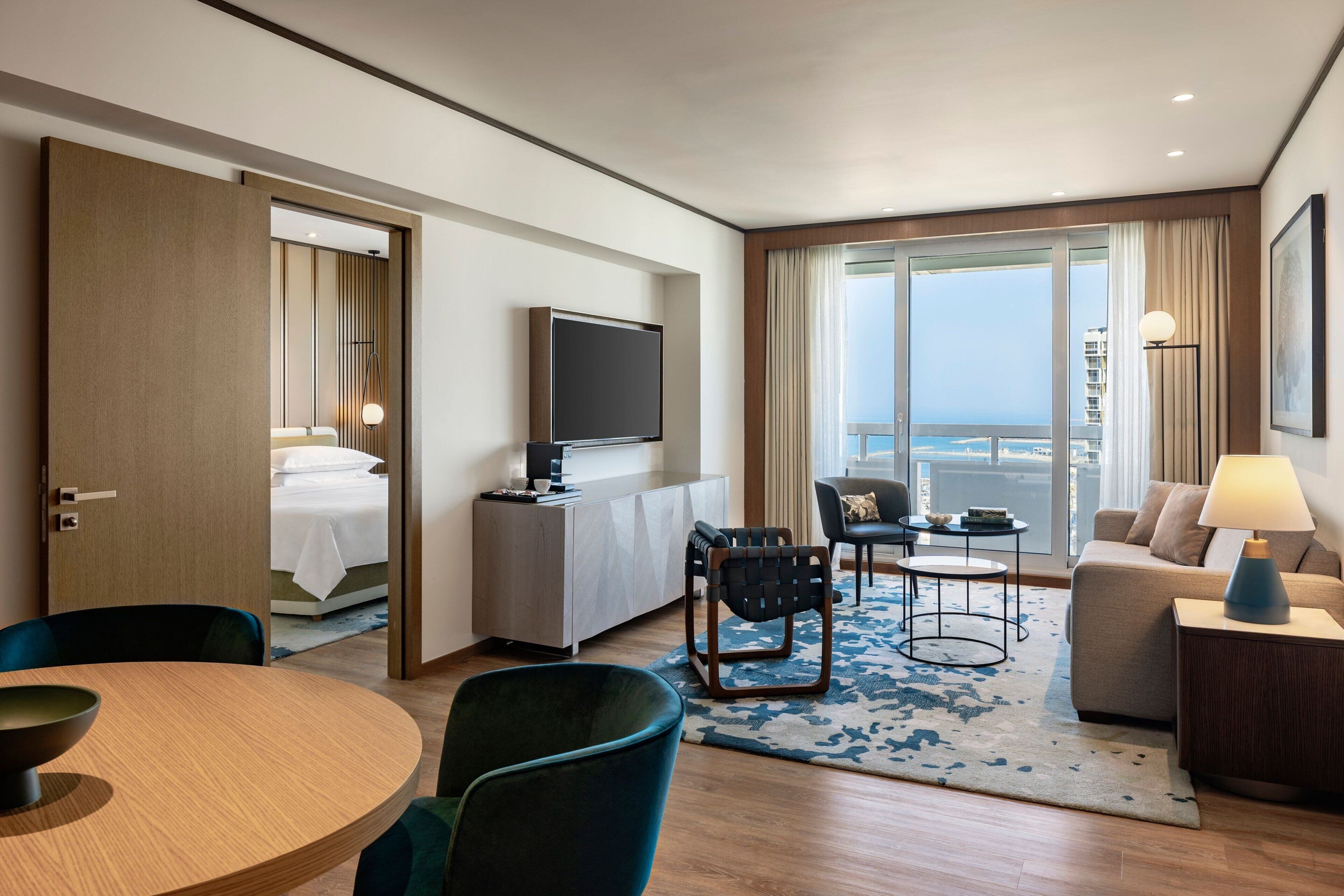 Habitación de hotel con cama, televisión, zona de comedor con sillas y mesa, sofá y vista al océano.