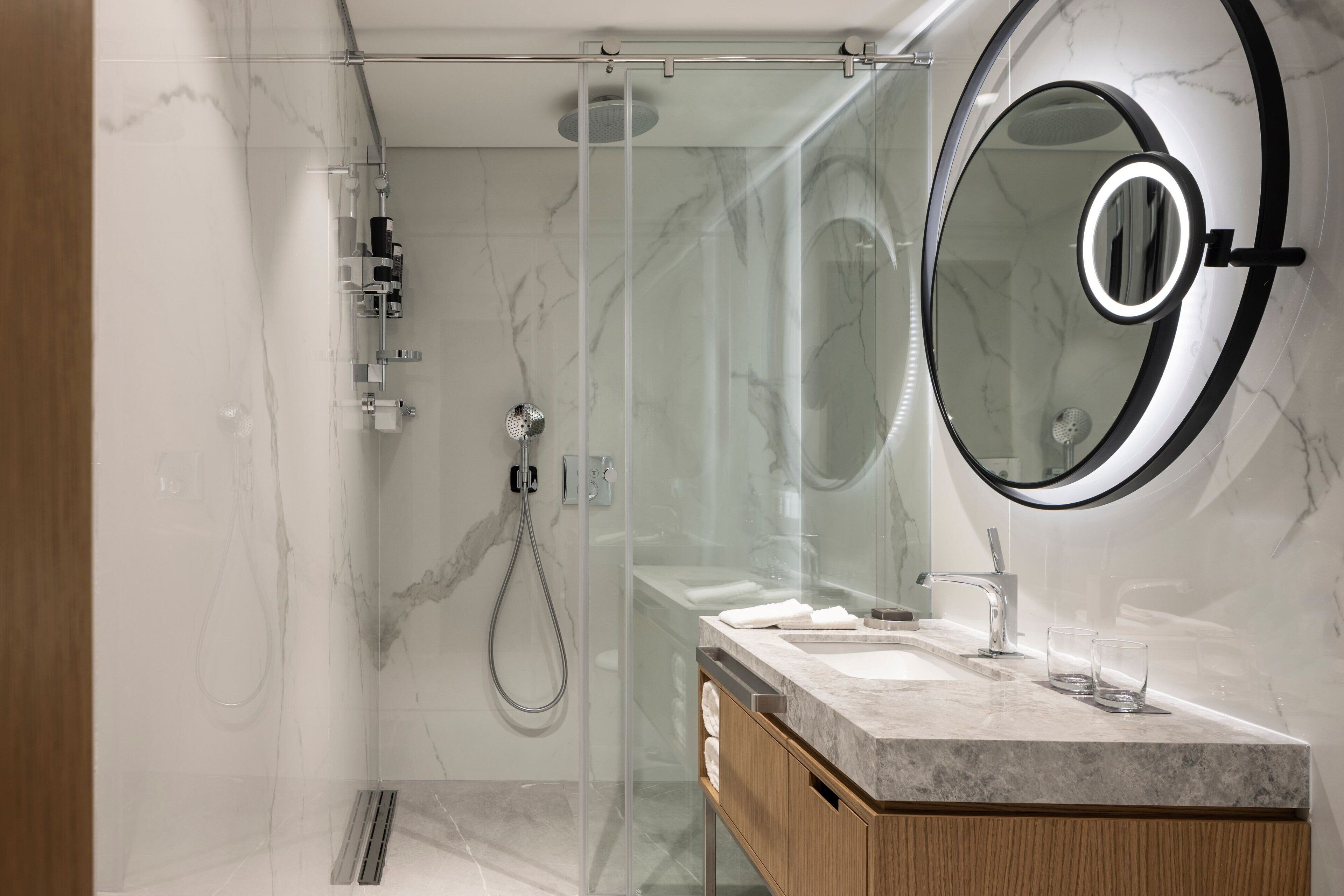 A modern bathroom with a glass-enclosed shower, a vanity with a marble countertop, and a round mirror with a light.