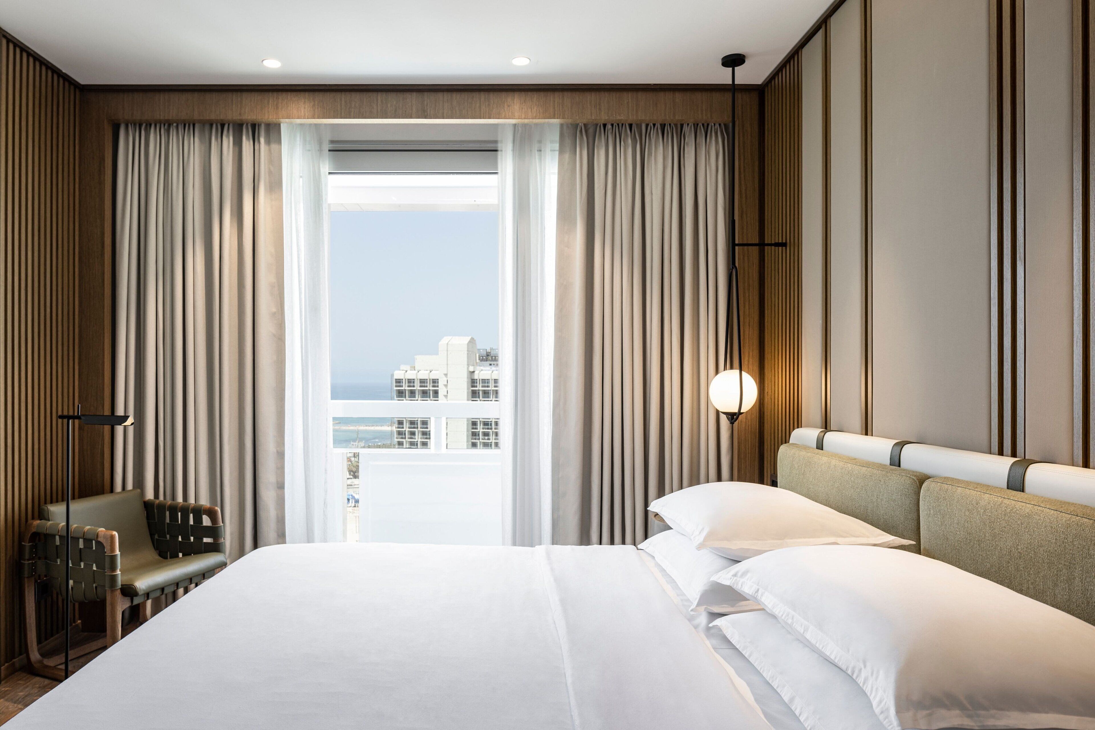 Une chambre d’hôtel avec un grand lit, une vue sur la ville par la fenêtre et un coin salon comprenant une chaise et une table.