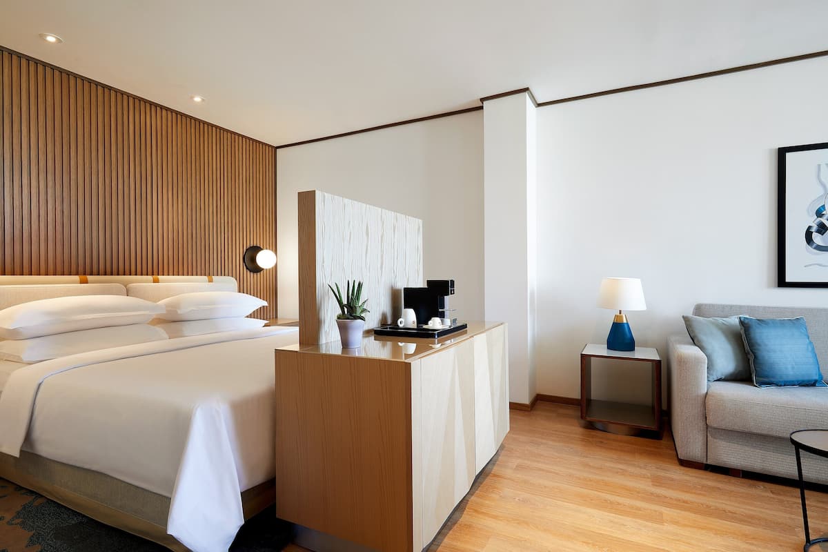 Una habitación de hotel moderna con una cama bien hecha, un escritorio pequeño con cafetera, un sofá y una lámpara.