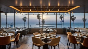 A restaurant with tables and chairs arranged for dining, large windows offering a view of the sea and palm trees, and hanging lights above the tables. - Sheraton Grand Tel Aviv (Tel Aviv)