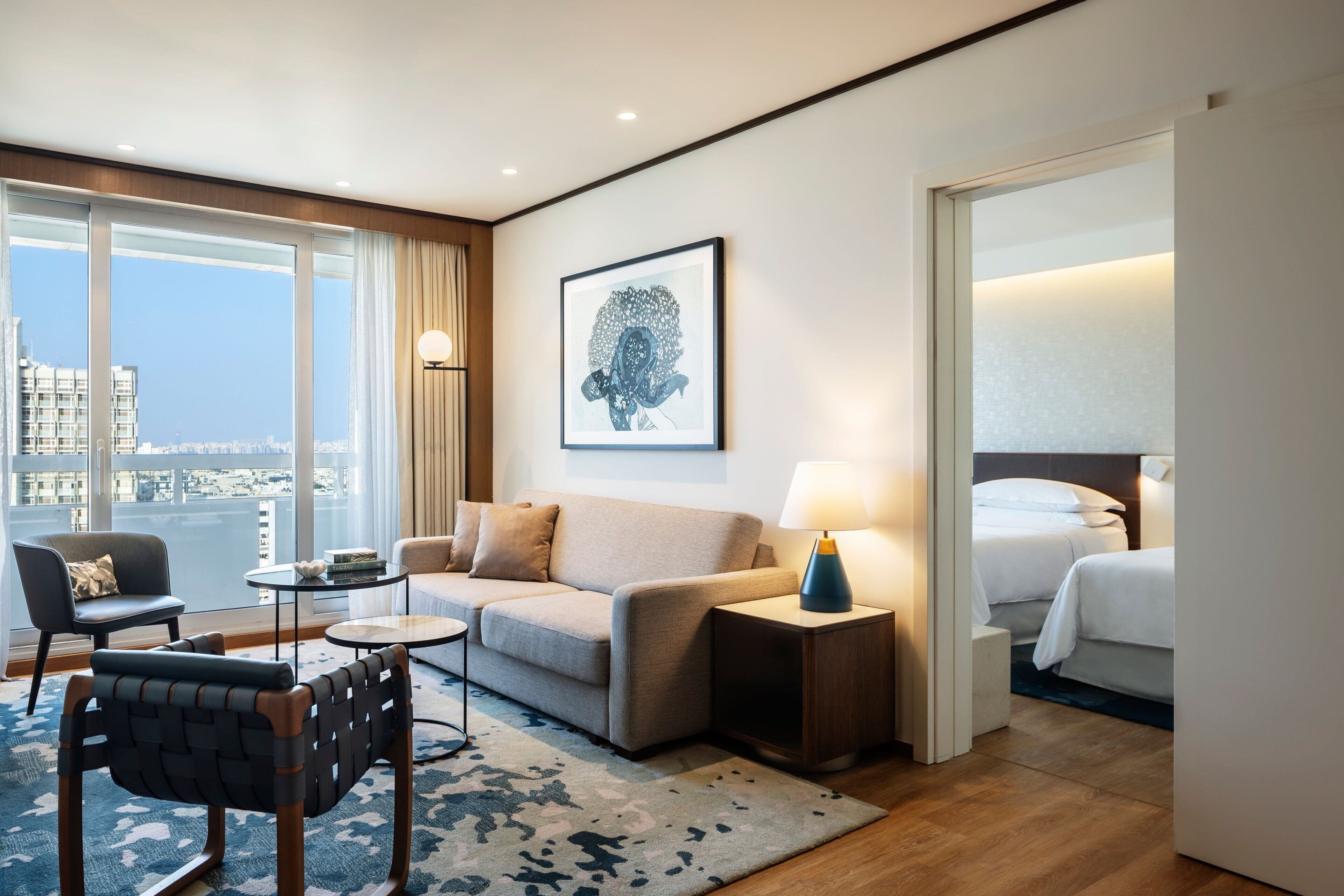 A modern hotel room with a large window, a sofa, a chair, a bedside table with a lamp, and a bed.