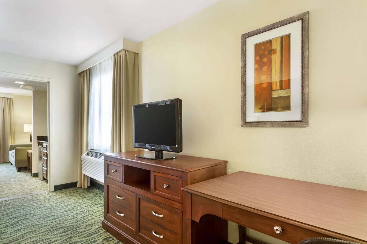 A hotel room with a wooden desk, a flat-screen TV, a framed picture on the wall, and a view of a hallway with a bed and a desk.