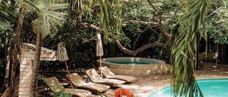 Zona de piscina con sillas de descanso y sombrillas, rodeada de exuberante vegetación.