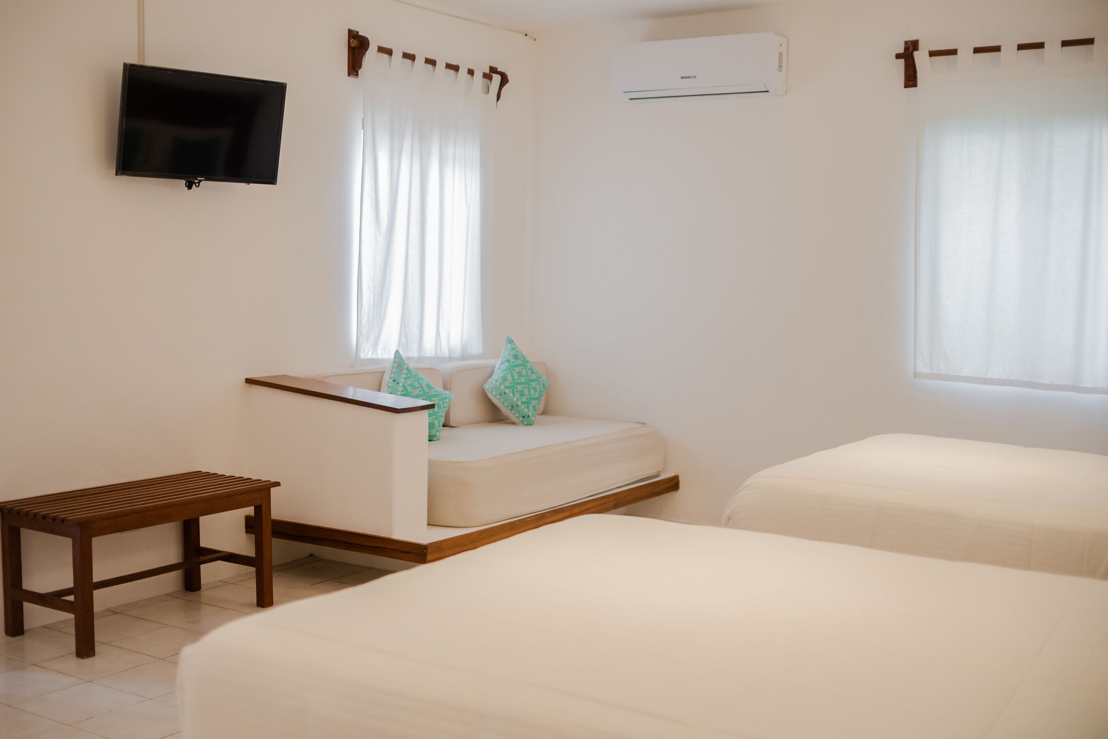 A hotel room with a bed, a small wooden table, a sofa with patterned cushions, a flat-screen TV mounted on the wall, and an air conditioning unit.