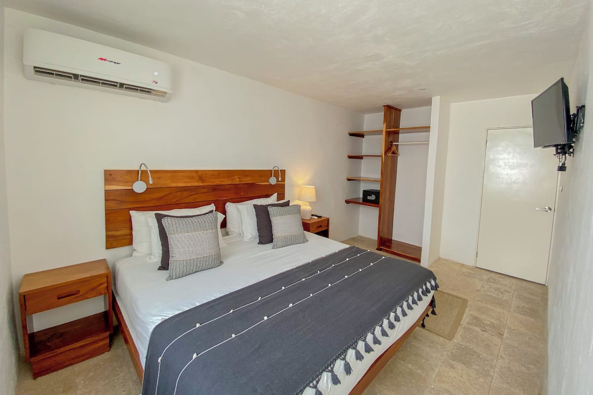 A bedroom with a wooden headboard, a bed with white and striped bedding, a nightstand, a wardrobe, and a television mounted on the wall.
