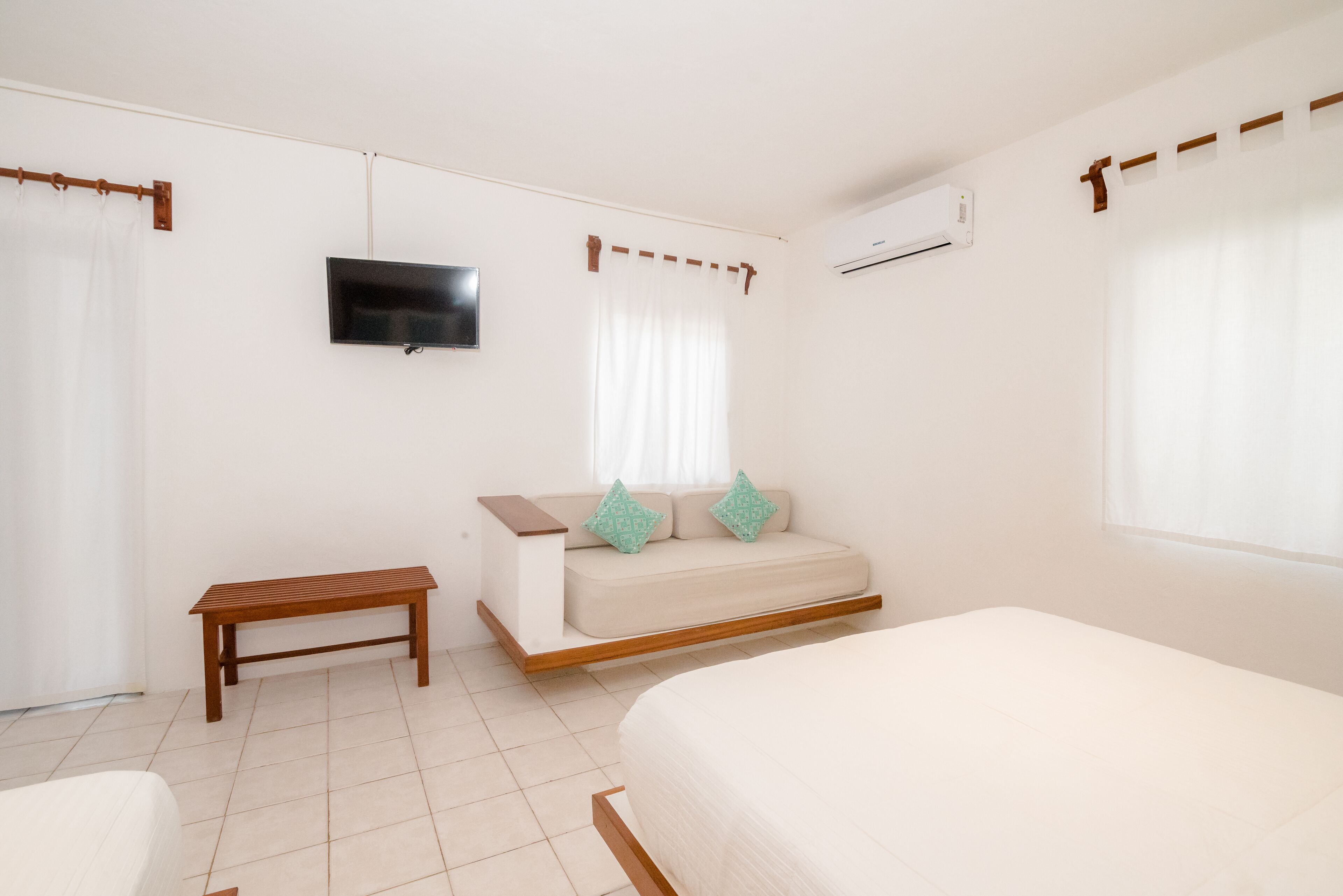 Un dormitorio con cama, una banca, un sofá con cojines, televisor y aire acondicionado.