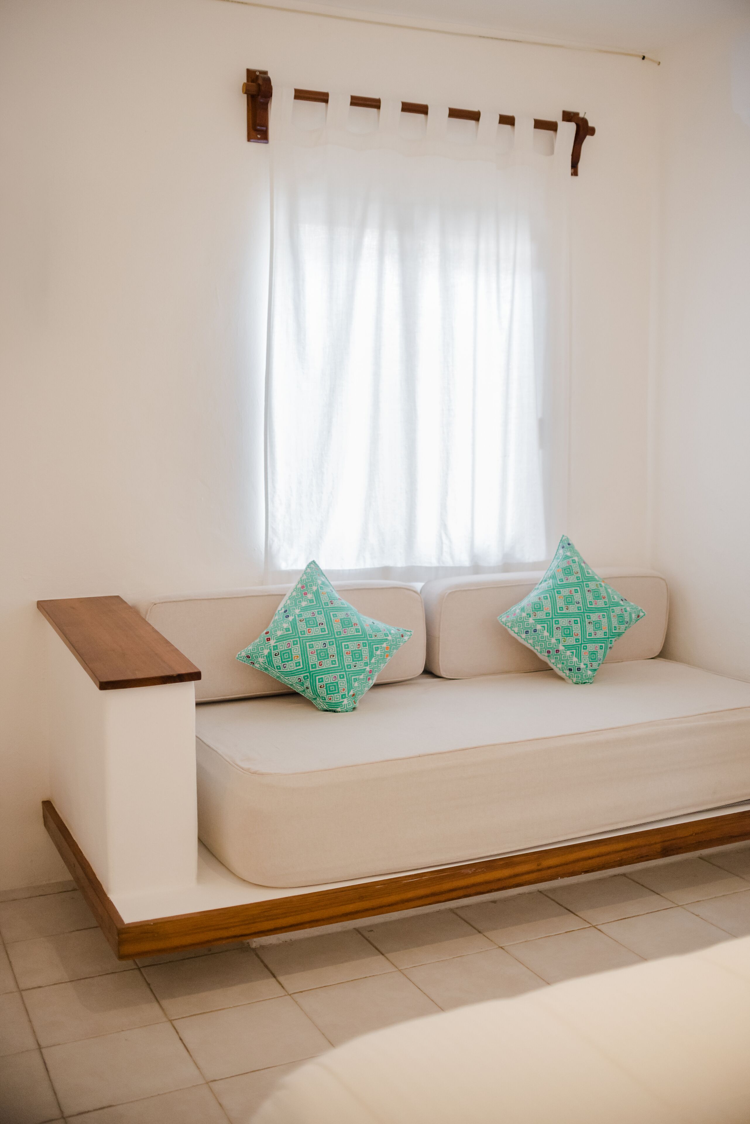A modern, minimalist room with a white sofa, two patterned cushions, a wooden side table, and a window with sheer curtains.