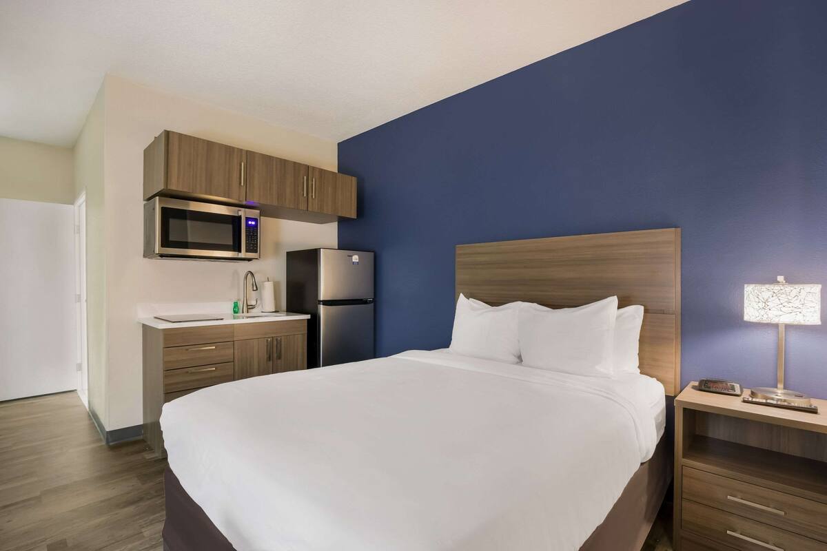 Una camera d'albergo con un letto, un comodino, una lampada, un forno a microonde, un frigorifero e un mobiletto in legno sopra il lavandino.