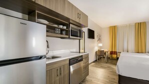 A compact hotel room with a kitchenette including a Galanz refrigerator, microwave, and dishwasher. There is a small dining area with a table and chairs, and a bed with a white comforter.