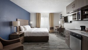 A hotel room with a bed, bedside table, lamp, chair, desk, and kitchenette.
