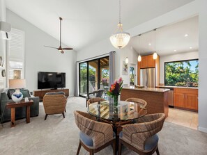 A modern living room with a TV, seating, a glass dining table, and a kitchen with wooden cabinets and a large window. - Wailea Ekahi - CoralTree Residence Collection (Kihei)