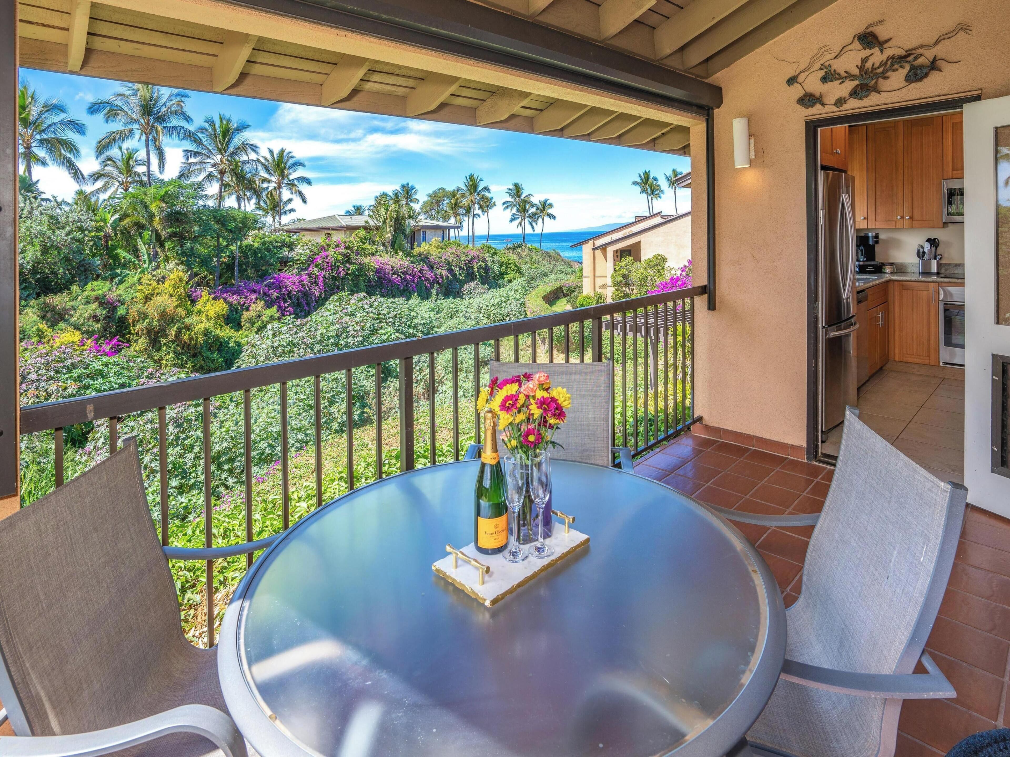 A balcony with a view of a tropical landscape, a table with a bottle and glasses, and a kitchen area.