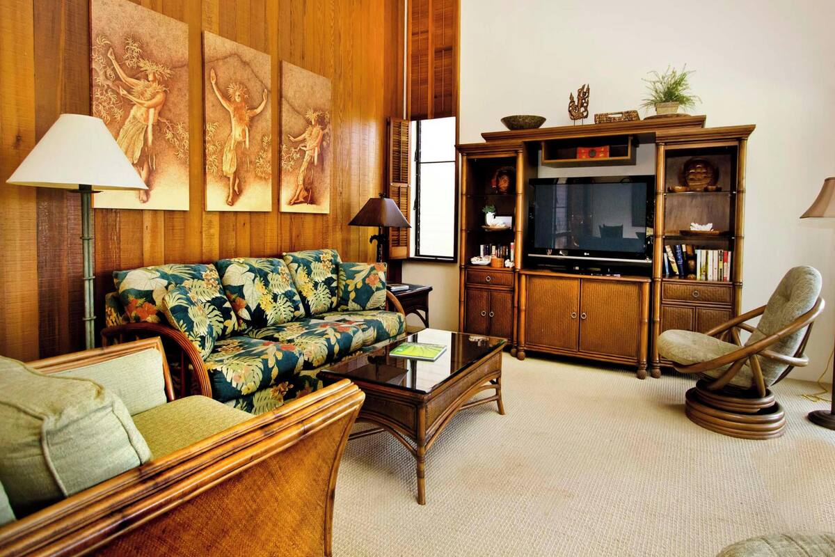 Un salón con sofá, sillones, una mesa de centro y televisor. Las paredes son de paneles de madera y hay una estantería con objetos decorativos.