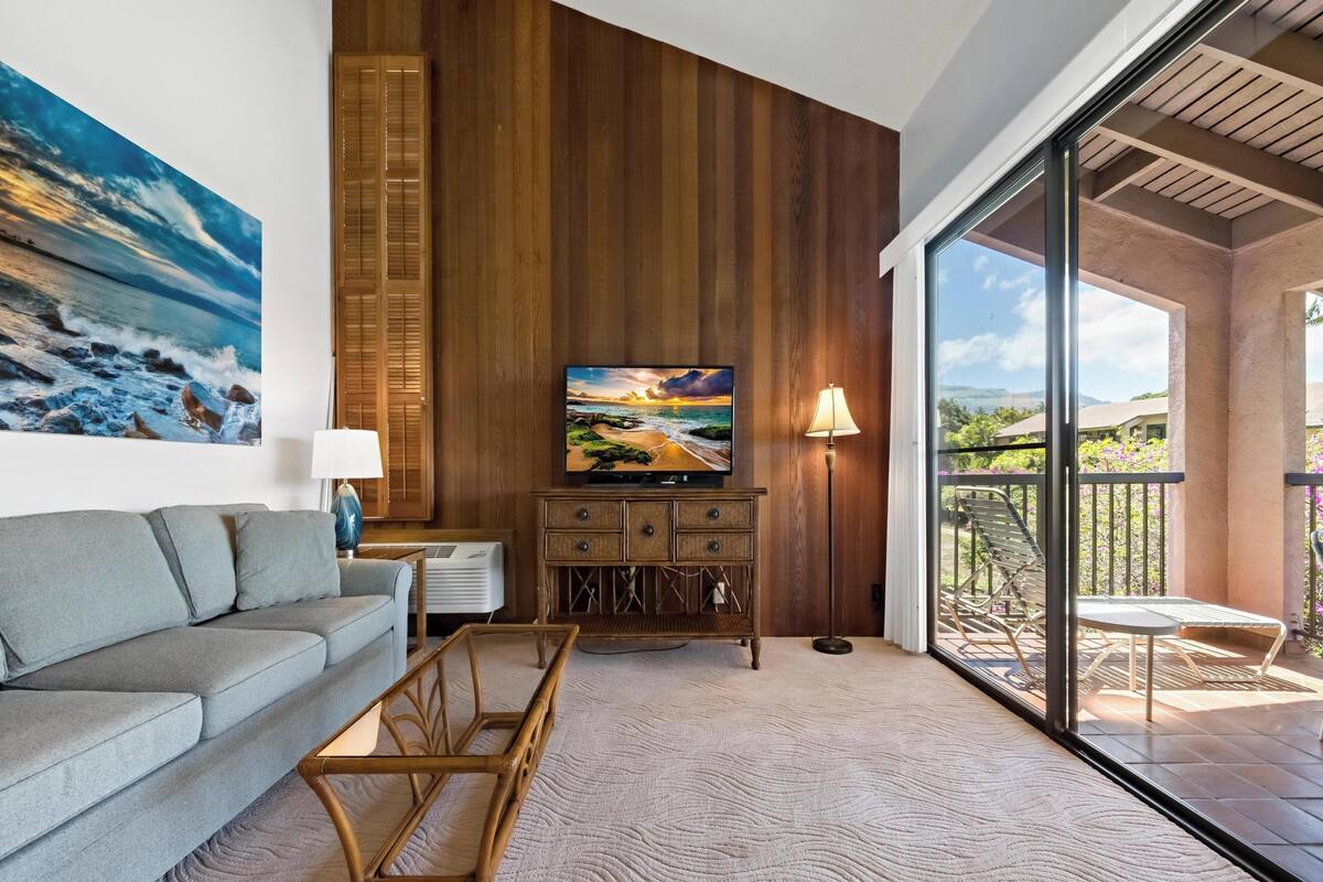 Una sala de estar con sofá, televisor, lámpara, aparador de madera y vista al exterior.
