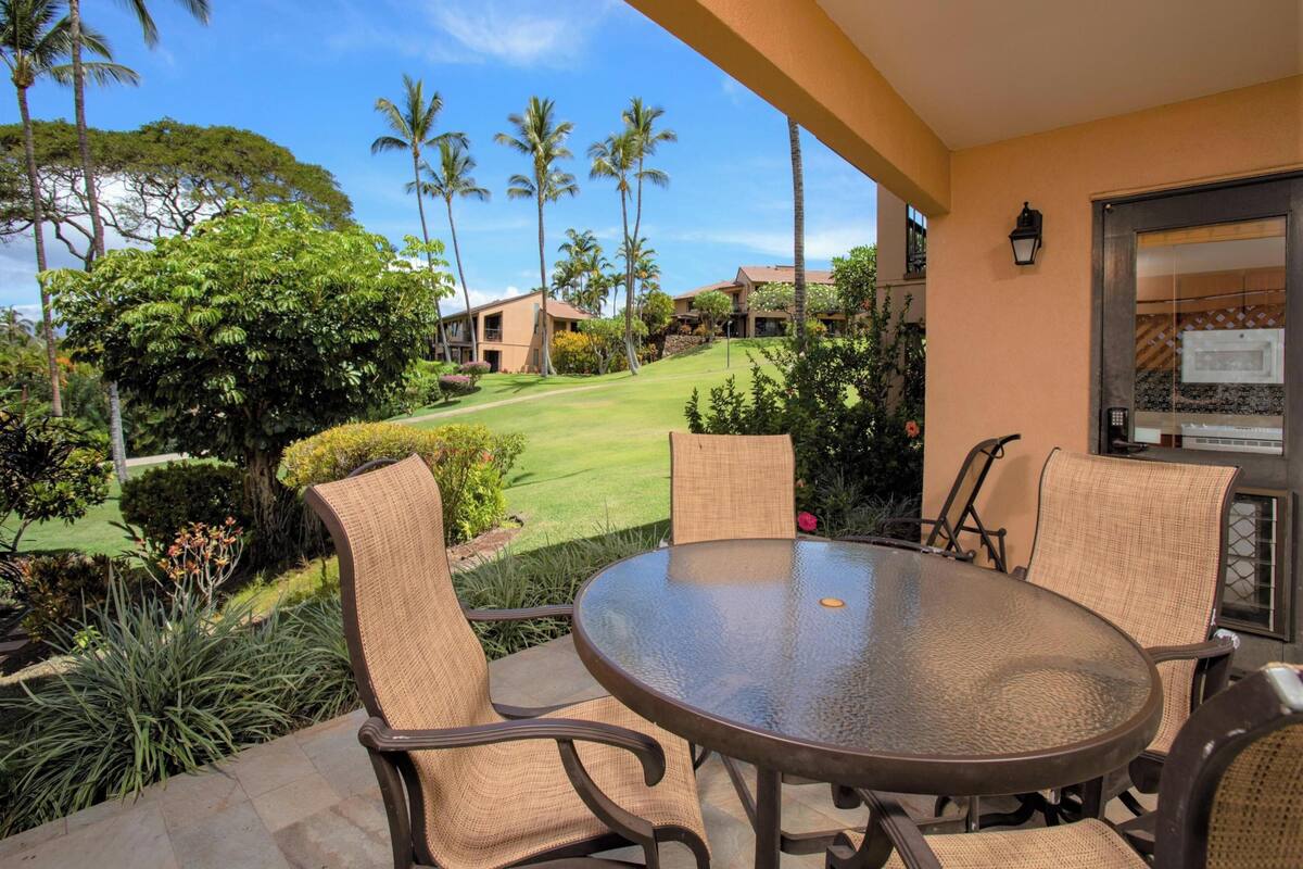 Un patio con una mesa redonda y sillas de mimbre, con vistas a un césped cuidado y palmeras.