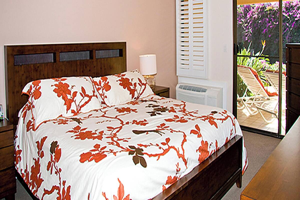 Un dormitorio con cama, mesita de noche, cómoda y vistas a un patio exterior con sillas y plantas.
