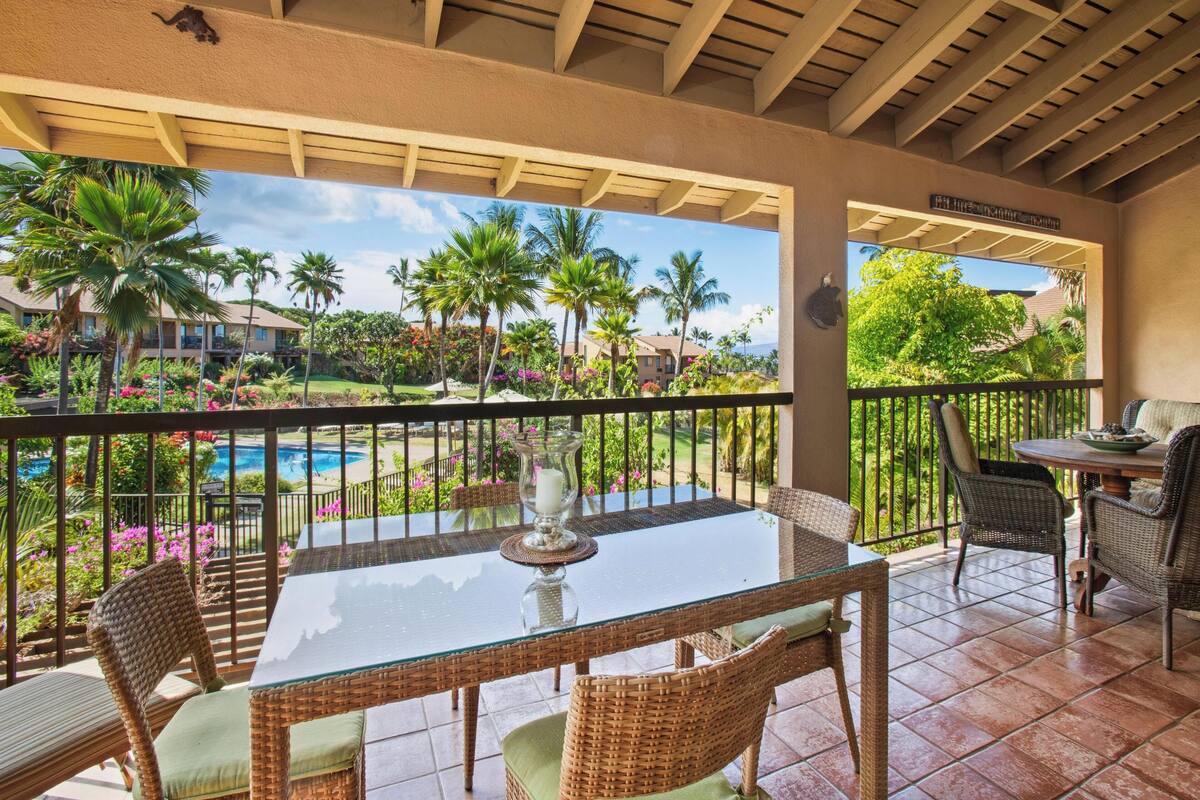 Un espacio exterior cubierto con una mesa de vidrio y sillas de mimbre, con vistas a una piscina y abundante vegetación.