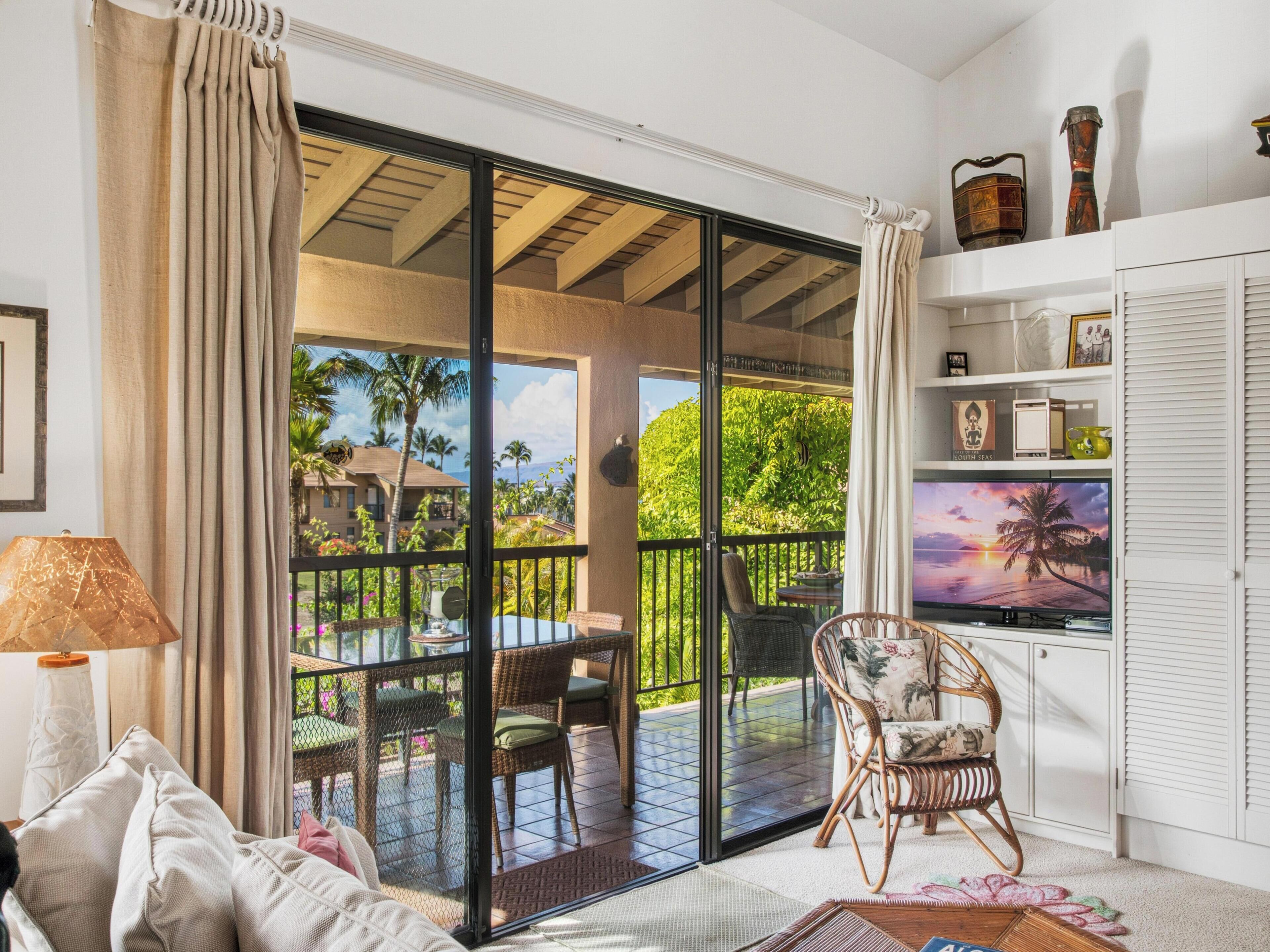A room with a sliding glass door leading to a balcony with outdoor furniture and a view of palm trees and buildings.