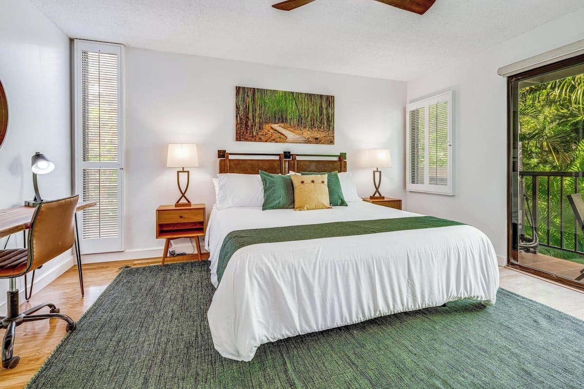 A bedroom with a bed, two bedside tables with lamps, a ceiling fan, a painting, and a window with blinds.