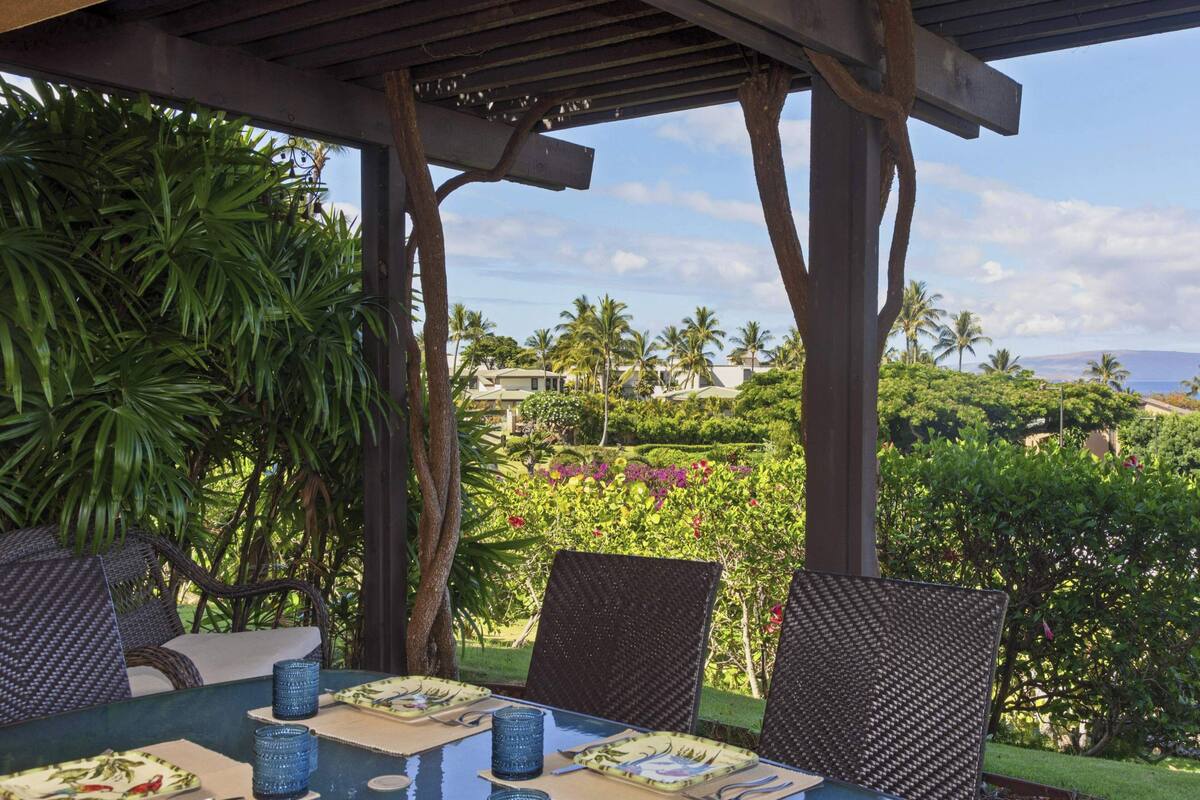 Un espacio de comedor al aire libre cubierto con una mesa para dos, rodeado de sillas de mimbre, con vistas a un jardín exuberante y al mar.