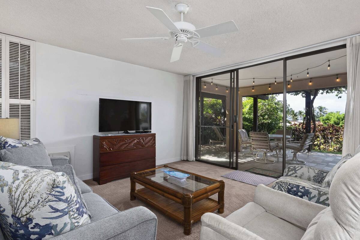 Sala de estar con una televisión de pantalla plana sobre un mueble de madera, ventilador de techo y un amplio ventanal corredizo que da a un patio exterior con mobiliario.