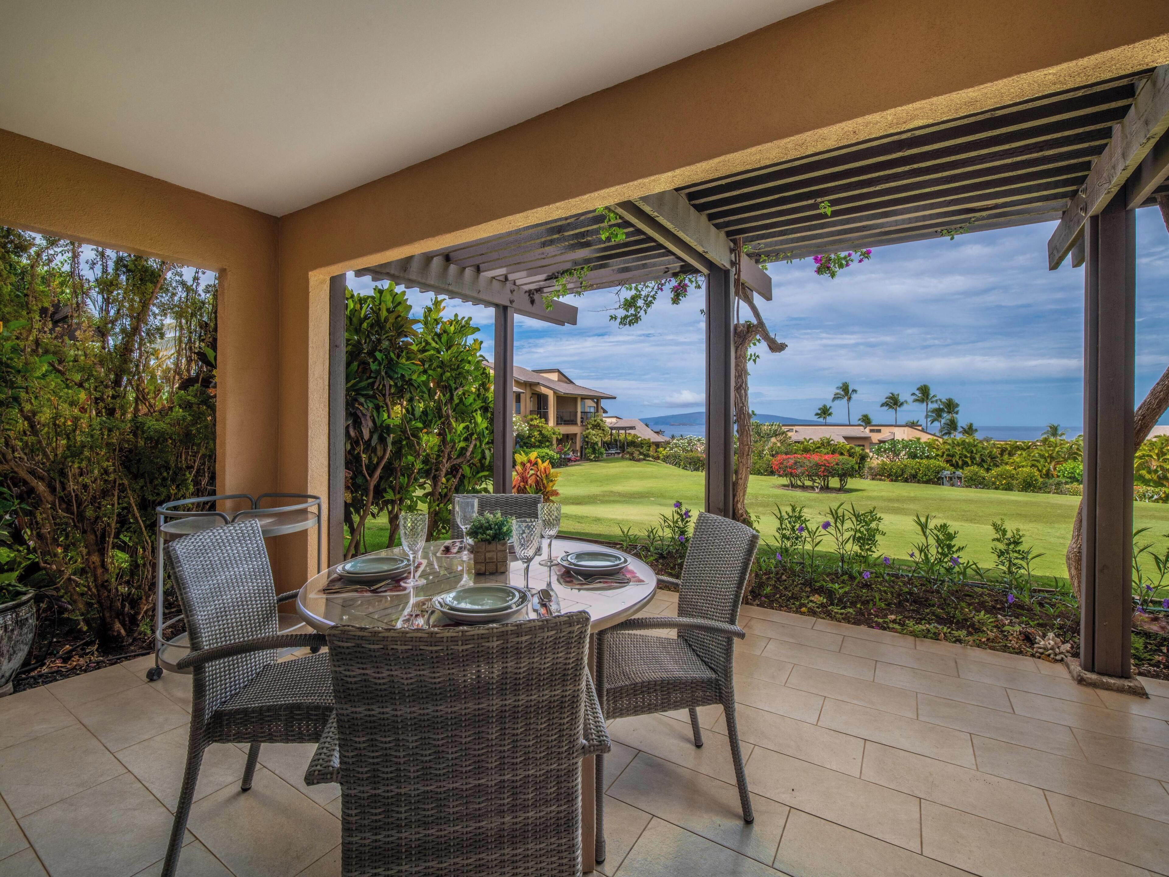 A covered outdoor dining area with wicker chairs and a table set for two, overlooking a well-manicured lawn and a view of the ocean.