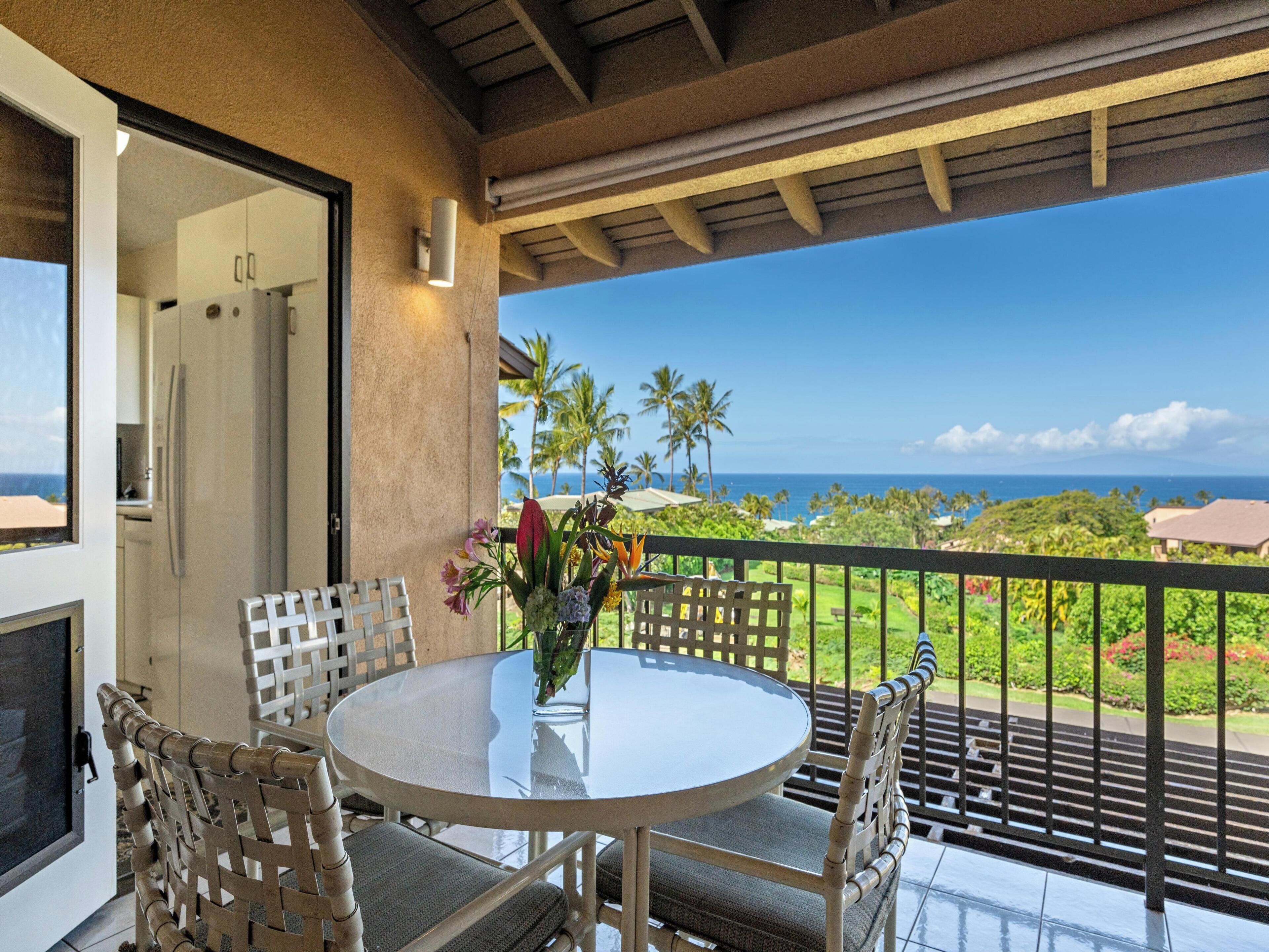 A balcony with a glass table and chairs, overlooking a lush landscape with palm trees and a view of the ocean.