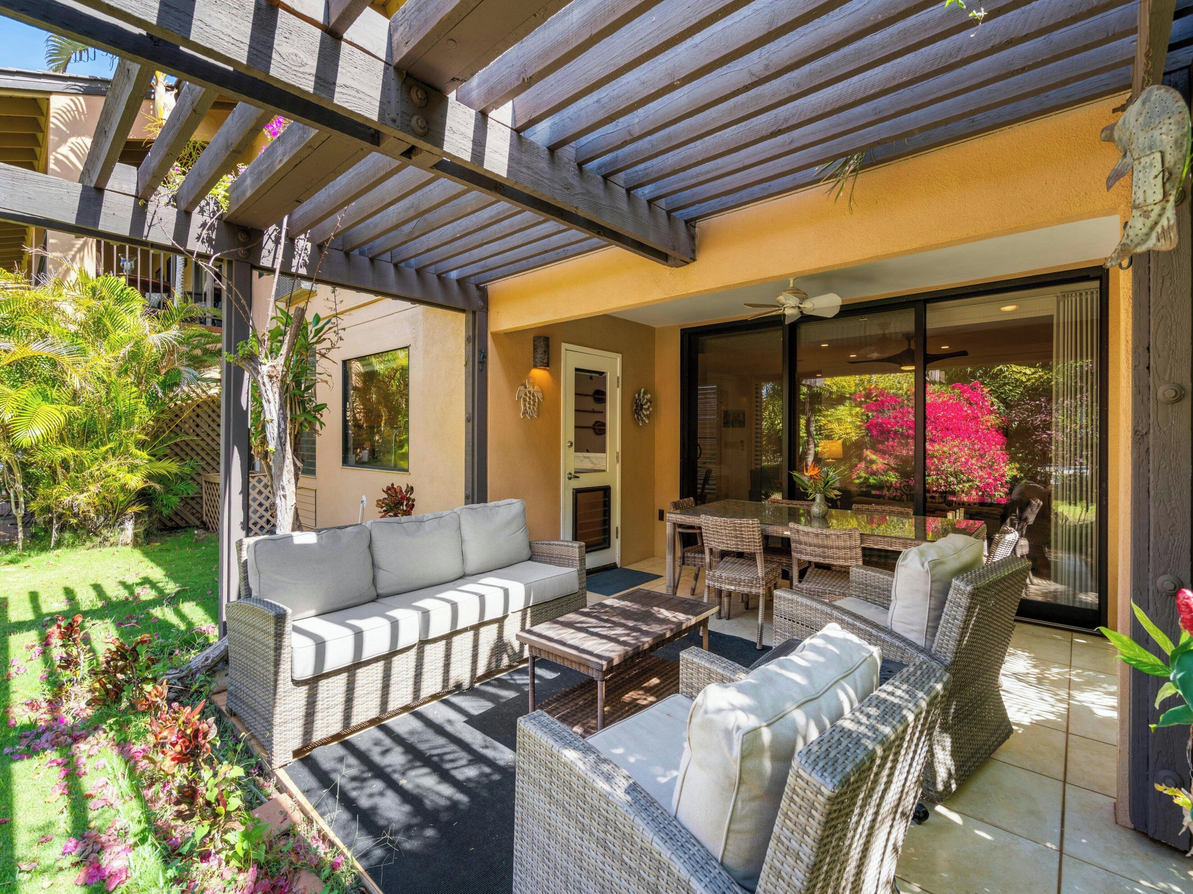 A covered patio area with wicker furniture, a glass table, and a view of a garden with pink flowers.